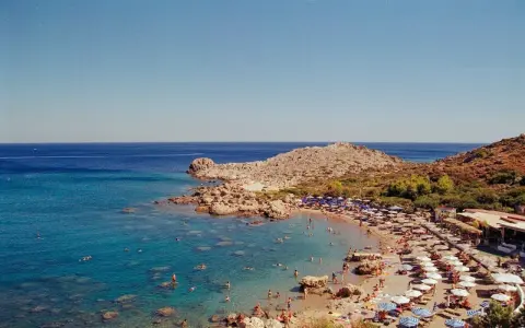 Rodos: turkusowe morze Egejskie, kamienista plaża z leżakami i parasolami, ludzie kąpiący się w wodzie.