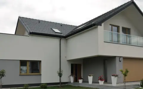 Nowoczesny dom z kremową elewacją, szarym cokołem, dużym oknem i balkonem ze szklaną balustradą. Przed wejściem białe donice z kwiatami.