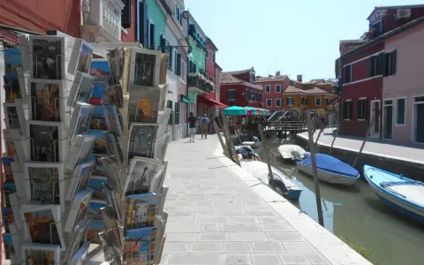 Cartoline di Murano, Burano e Torcello in vendita lungo un canale con case colorate e barche ormeggiate.