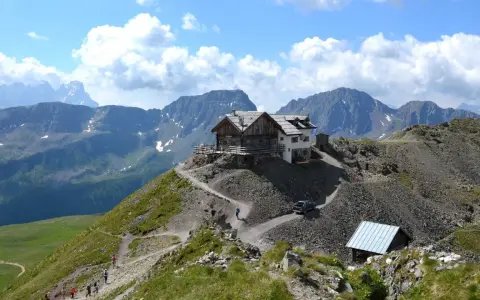 Rifugio alpino su un crinale roccioso, con sentiero tortuoso e escursionisti. Un'auto si avvicina al passo stalle.