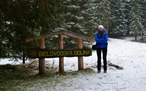 Kobieta w niebieskiej kurtce stoi przy drewnianym znaku "Bielovodská dolina" w zimowej scenerii.