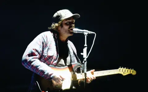 Neil Halstead, con gorra y camisa a cuadros, toca la guitarra y canta en un concierto.