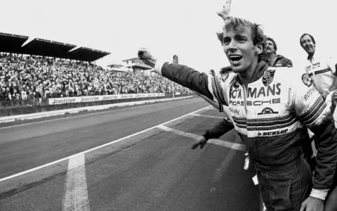 Jubelnder Stefan Bellof im Rennanzug mit "Le Mans" und "Porsche" Aufschrift, jubelnde Menge im Hintergrund.