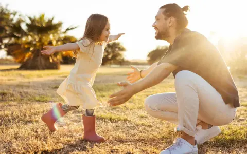Ein kleines Mädchen rennt mit offenen Armen auf ihren lachenden Vater zu. Die warme Abendsonne beleuchtet die Szene und unterstreicht die liebevolle vater tochter beziehung.
