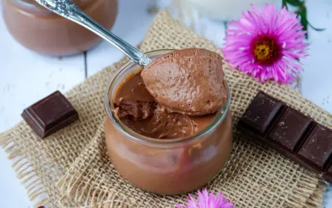 Une cuillère de crème au chocolat sans sucre, onctueuse et aérienne, sur un pot en verre. Des carrés de chocolat et des fleurs roses décorent la scène.