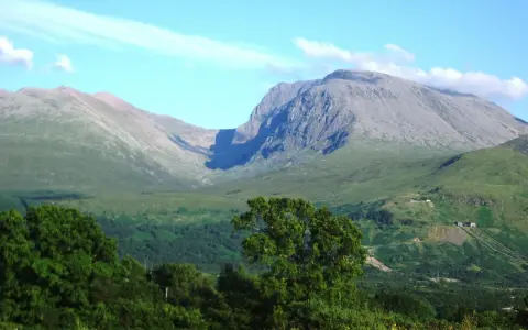 Majestatyczny Ben Nevis wznosi się na tle błękitnego nieba z delikatnymi chmurami. Zielone zbocza górskie i bujna roślinność tworzą malowniczy krajobraz.