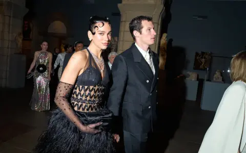 Dua Lipa stuns at the Met Gala in a black feathered gown and diamond necklace, walking with a companion.