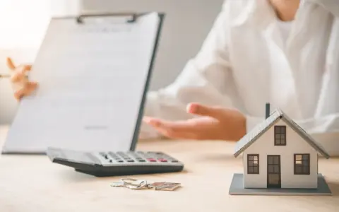 Une personne présente un contrat, une calculatrice, des clés et une maquette de maison. L'ambiance est un peu sinistre, comme si un problème était sur le point d'être révélé.