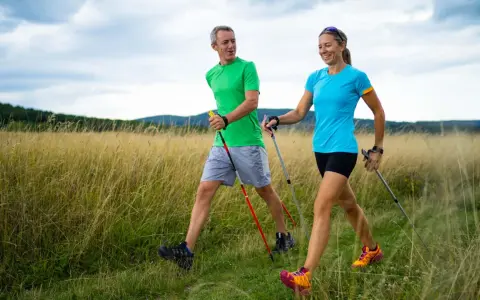 Ein Paar genießt das **power walking** in einer sonnigen Landschaft mit hohem Gras und bewaldeten Hügeln im Hintergrund.