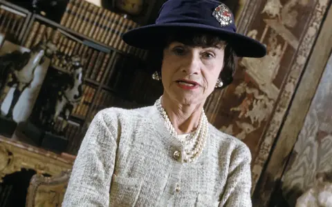 Iconica stilista Coco Chanel, con cappello blu e perle, in un ambiente elegante con libri e decorazioni.