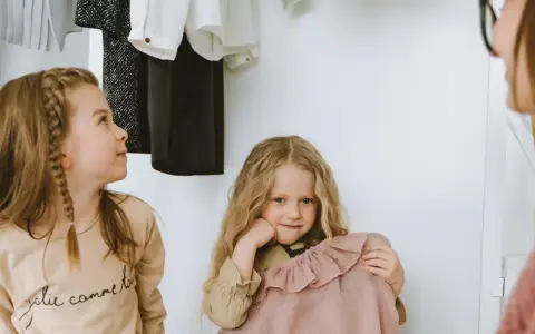 Dwie dziewczynki wybierają ubrania z szafy. Jedna ma warkocz, druga trzyma różową sukienkę.