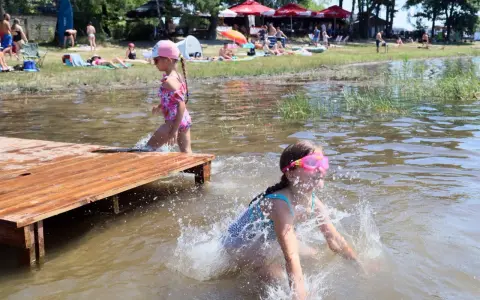 Dwie dziewczynki pluskają się w wodzie przy drewnianym pomoście. W tle plażowicze i drzewa. Wakacje nad jeziora lubelskie.