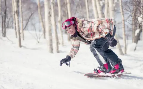 Uśmiechnięta snowboardzistka z różowymi włosami zjeżdża po ośnieżonym stoku wśród drzew. To świetna zabawa podczas chodzenia po górach zimą.
