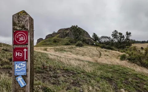 Wanderwegweiser mit Falken-Symbol und "H2"-Markierung, der zu den **rundwanderwege owl** führt. Ein Pfad schlängelt sich über einen grasbewachsenen Hügel zu einer Felsformation.