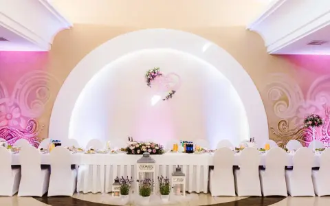 Elegancka sala weselna z białymi pokrowcami na krzesłach i serwowanym stołem. Serce z imionami pary młodej zdobi ścianę, tworząc atmosferę wesela z klasą.