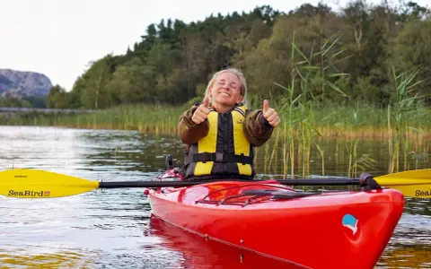 Glad kvinna i kajak med all kajak utrustning, ger tummen upp. Vacker natur med vass och träd i bakgrunden.