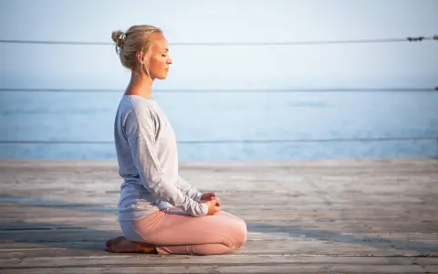 Kvinna med slutna ögon sitter i meditationsställning på en träbrygga vid havet, utövar progressiv avslappning.