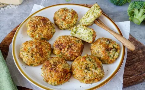 Goldbraune, knusprige broccoli nuggets auf einem Teller, ein Stück angeschnitten, mit frischem Brokkoli im Hintergrund.