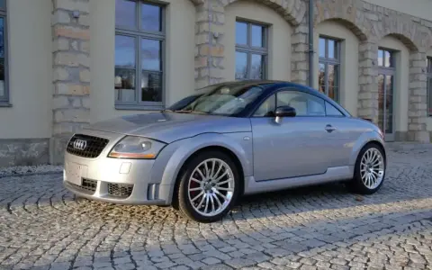 Silberner Audi TT 2005 mit schwarzem Dach parkt auf Kopfsteinpflaster vor einem Gebäude mit Steinbögen.