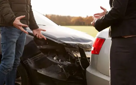 Dwie osoby dyskutują przy uszkodzonych samochodach po kolizji. Mężczyzna gestykuluje, wskazując na zniszczony przód czarnego auta. Kobieta stoi przy srebrnym samochodzie, również wyrażając swoje emocje. Trudno ustalić numer polisy OC bez dokumentów.