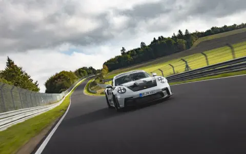 Biały Porsche 911 GT3 RS z czarnymi akcentami pędzi po torze wyścigowym wśród zielonych wzgórz i chmur.