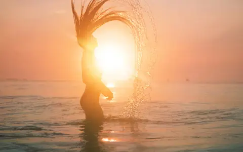 Silhouette d'une femme jetant ses cheveux dans l'eau au coucher du soleil, avec un effet de lens flare photoshop.