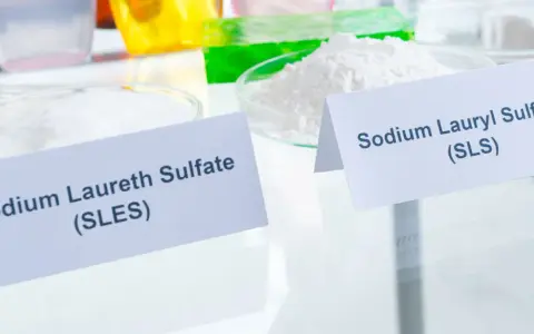 Zwei Schalen mit weißen Pulvern, beschriftet mit "Sodium Laureth Sulfate (SLES)" und "Sodium Lauryl Sulfate (SLS)". Diese Inhaltsstoffe sind oft in SLS-Shampoos zu finden.