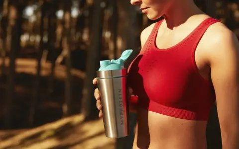 En kvinna i röd sporttopp håller en proteinshaker. Frågan om hur mycket protein kan kroppen ta upp är relevant för hennes träning.