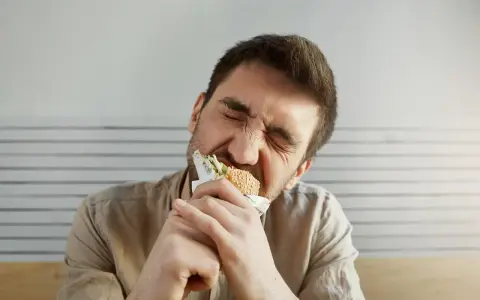 Uomo con espressione sofferente mangia un panino, forse per alleviare lo stress, ma teme di ingrassare.