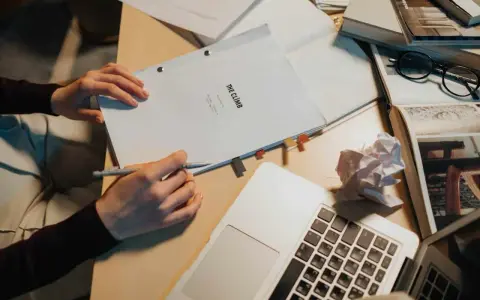 Manos escribiendo un guion cortometraje, rodeado de libros, gafas y un portátil.