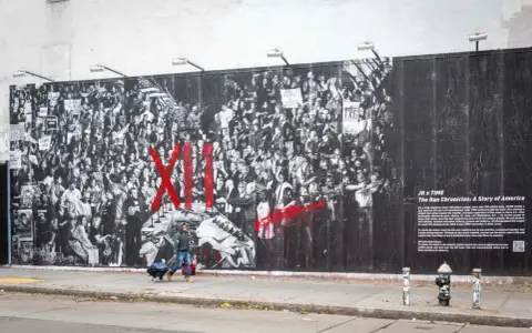 Une fresque murale de street art d'origine, "The Gun Chronicles: A Story of America", par JR x TIME, dépeint une foule en noir et blanc avec des touches de rouge vif.