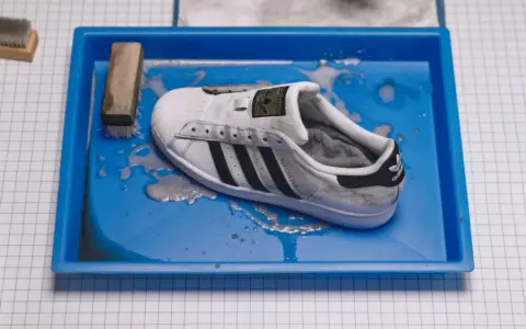Ein schmutziger weißer Adidas Superstar Schuh liegt in einer blauen Wanne mit Wasser und Seife. Reinigungswerkzeuge liegen daneben.
