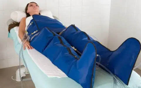 Kobieta leży na łóżku, jej nogi są owinięte w niebieskie mankiety urządzenia do mechanicznego drenażu limfatycznego.