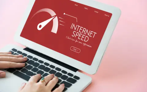 Schermo di un laptop con indicatore di velocità internet su sfondo rosso. Le mani digitano sulla tastiera, frustrati dall'internet lento.