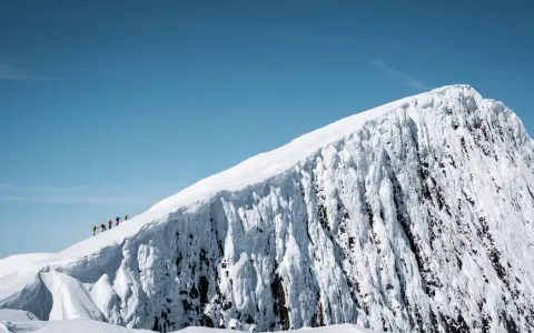 En grupp vandrare på en snöig bergstopp under en helags topptur. Den isiga bergväggen reser sig dramatiskt mot en klarblå himmel.