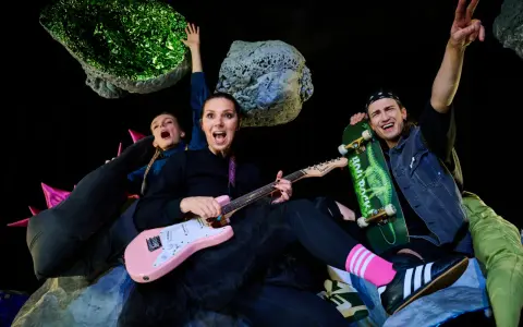 Energiczna ekipa w kosmicznej scenografii, grająca polski folk rock. Kobieta z różową gitarą, mężczyzna ze skateboardem, wesołe miny i podniesione ręce.