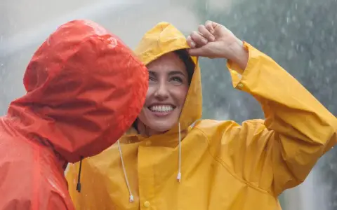 Zwei lachende Personen im Regen, eine in Gelb, die andere in Rot. Die gelbe Regenjacke ist im Fokus.