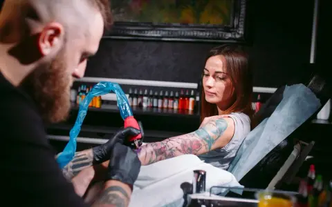 Artysta tworzy świeży tatuaż na ramieniu kobiety. Po sesji będzie musiała unikać pracy fizycznej, by skóra mogła się zagoić.