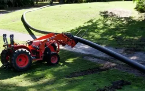 A tractor with a front loader is laying a long, flexible HDPE pipe across a grassy area.