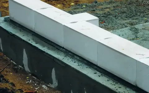 Białe bloczki betonowe układane na warstwie izolacji. Widać wilgotną zaprawę i ziemię wokół fundamentów.