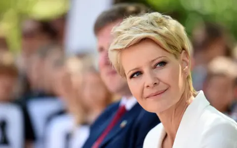 Kobieta z blond włosami w super krótkiej fryzurze damskiej, w białej marynarce, uśmiecha się.