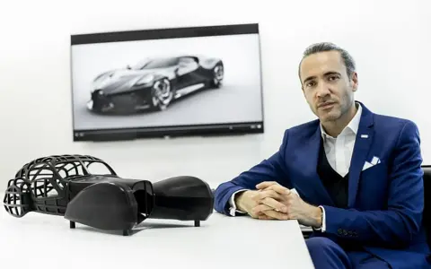 Ein Mann in einem blauen Anzug sitzt vor einem Bildschirm mit dem Bugatti La Voiture Noire. Daneben ein Modell des Autos.
