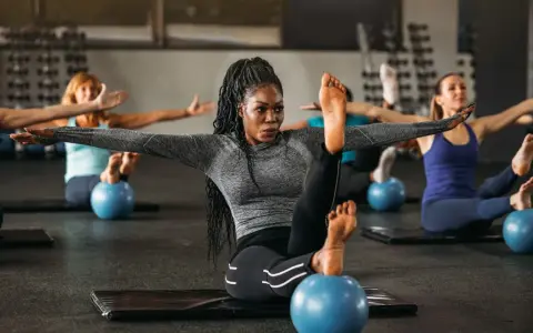 Frauen in einem Kurs, der Pilates Krafttraining macht, sitzen auf Matten mit kleinen blauen Bällen unter ihren Füßen.