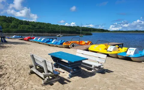 Słoneczny dzień nad jeziorem Sarbsko. Rząd kolorowych rowerów wodnych czeka na plaży, obok drewniane ławki i stół.