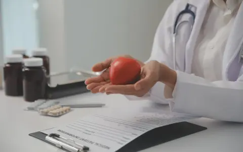 Lekarz trzyma w dłoniach czerwone serce, symbolizujące zdrowie. Na stole leżą leki i dokumentacja medyczna, sugerując rozmowę o tym, po jakim czasie spada cholesterol.