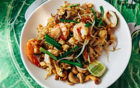 Talerz Pad Thai z krewetkami, kalmarami, kiełkami i orzechami nerkowca, udekorowany ćwiartką limonki.