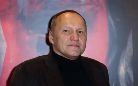 Portret Mariusza Grzegorzka w czarnym golfie i brązowej marynarce na tle abstrakcyjnego wzoru.