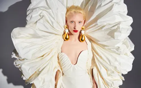 A model wears a dramatic white gown with a voluminous, ruffled cape, showcasing Daniel Roseberry's avant-garde design and striking gold teardrop earrings.