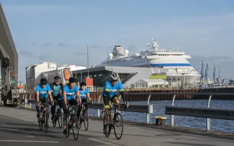 Fünf Radfahrer in blauen Trikots trainieren für einen Mini-Triathlon am Hafen, ein großes Kreuzfahrtschiff im Hintergrund.