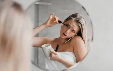 Une femme, enroulée dans une serviette, applique son mascara tout en tenant une tasse et en parlant au téléphone. Le mascara qui coule sur son visage témoigne de sa matinée mouvementée.
